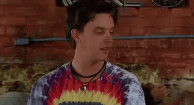 A Man Wearing A Tie Dye Shirt And A Choker Is Sitting On A Couch In Front Of A Brick Wall . GIF