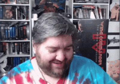 A Man Wearing A Tie Dye Shirt Is Smiling In Front Of A Book Titled Wing Commander GIF