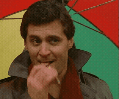 A Man Wearing A Trench Coat And Scarf Is Biting A Cigarette While Holding A Colorful Umbrella GIF