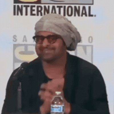 A Man Wearing A Turban And Glasses Is Holding A Bottle Of Water . GIF