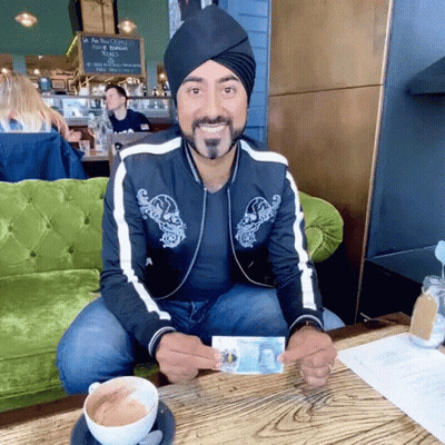 A Man Wearing A Turban Is Sitting At A Table Holding A Dollar Bill GIF
