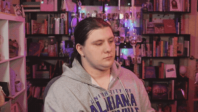 A Man Wearing A University Of Alabama Sweatshirt Sits In Front Of A Bookshelf GIF