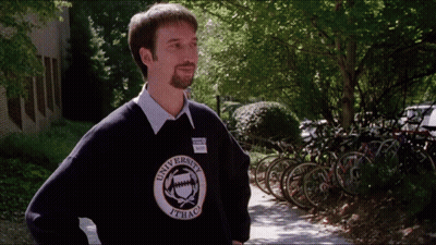 A Man Wearing A University Of Ithaca Sweater Stands On A Sidewalk GIF