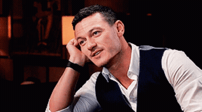 A Man Wearing A Vest And A White Shirt Is Sitting Down With His Hand On His Chin . GIF