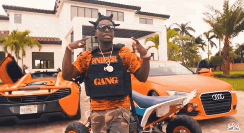 A Man Wearing A Vest That Says Gang Is Standing In Front Of Two Orange Cars GIF