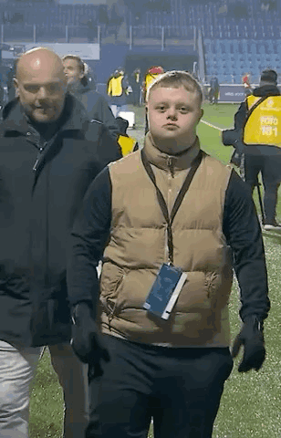 A Man Wearing A Vest With A Lanyard Around His Neck Is Walking On A Field . GIF