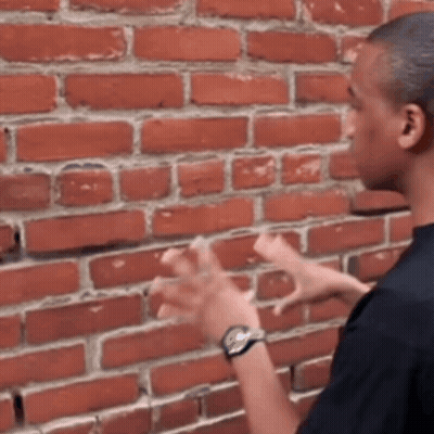 A Man Wearing A Watch Is Standing In Front Of A Brick Wall With His Hands In The Air . GIF