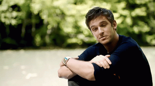 A Man Wearing A Watch Sits With His Arms Crossed In Front Of Trees GIF