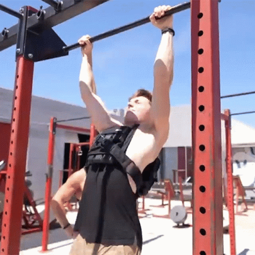 A Man Wearing A Weight Vest Hangs From A Bar In A Gym GIF