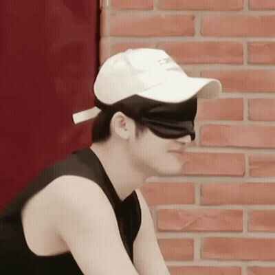 A Man Wearing A White Baseball Cap And Blindfold Is Sitting In Front Of A Brick Wall . GIF