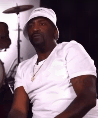 A Man Wearing A White Bucket Hat And A White Shirt Is Sitting In Front Of A Drum Set . GIF