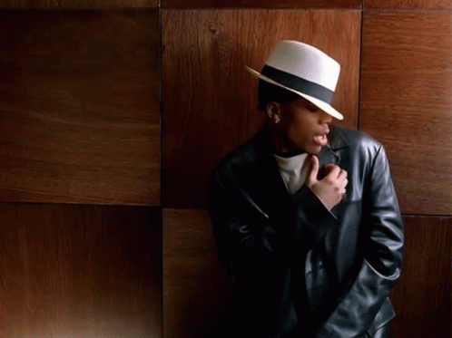 A Man Wearing A White Hat And A Black Leather Jacket Stands In Front Of A Wooden Wall GIF