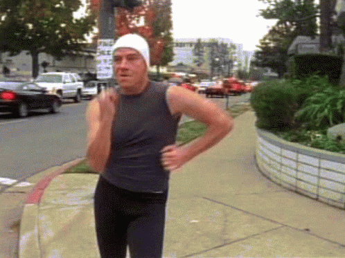 A Man Wearing A White Hat And A Black Tank Top Is Running Down A Sidewalk GIF