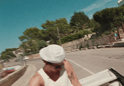 A Man Wearing A White Hat And A White Tank Top Is Walking Down A Street GIF