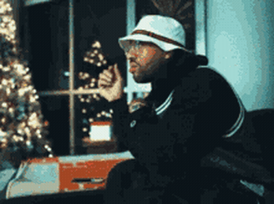 A Man Wearing A White Hat And Glasses Is Sitting In Front Of A Christmas Tree . GIF