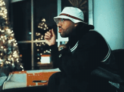 A Man Wearing A White Hat And Glasses Sitting In Front Of A Christmas Tree GIF