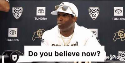 A Man Wearing A White Hat And Sunglasses Is Sitting In Front Of A Sign That Says " Do You Believe Now " GIF