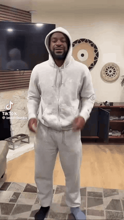 A Man Wearing A White Hoodie And Sweatpants Is Standing In A Living Room . GIF
