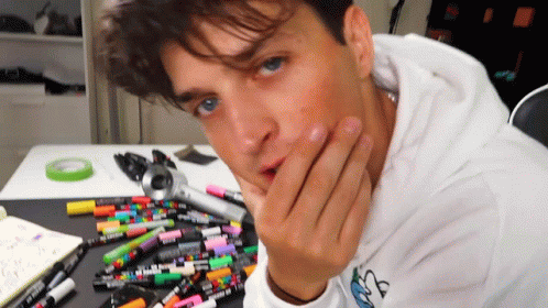 A Man Wearing A White Hoodie With A Blue Butterfly On It Is Sitting At A Table Full Of Markers GIF