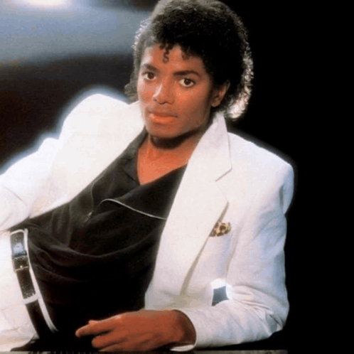 A Man Wearing A White Jacket And A Black Shirt Is Sitting Down GIF