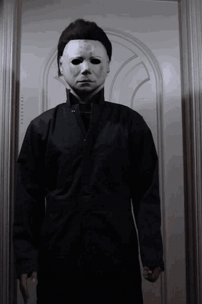 A Man Wearing A White Mask And A Black Jacket Is Standing In Front Of A Door GIF