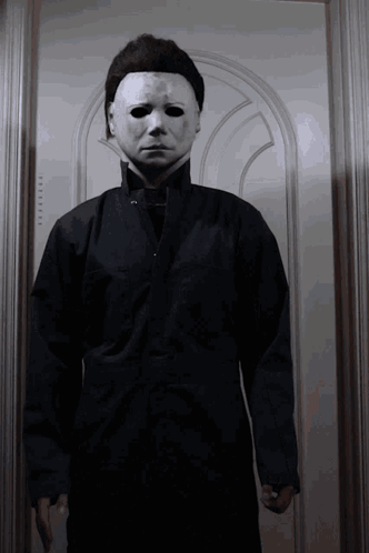 A Man Wearing A White Mask And A Black Jacket Is Standing In Front Of A Door GIF