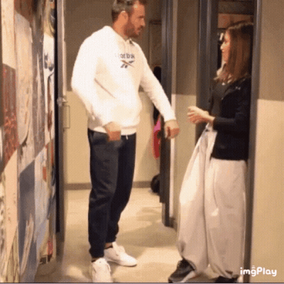 A Man Wearing A White Reebok Sweatshirt Talks To A Woman In A Hallway GIF
