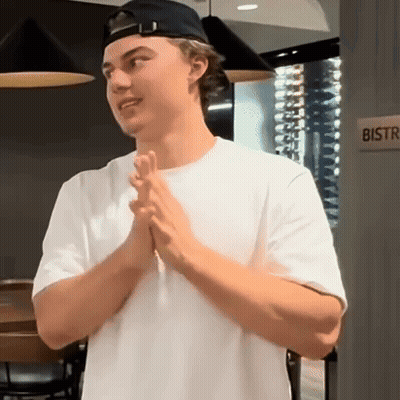 A Man Wearing A White Shirt And A Black Hat Is Standing In Front Of A Bistro Sign . GIF