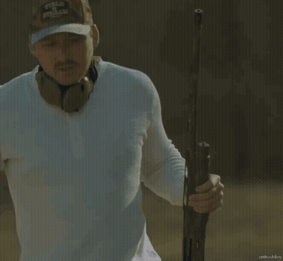 A Man Wearing A White Shirt And A Camo Hat Holds A Gun In His Right Hand GIF