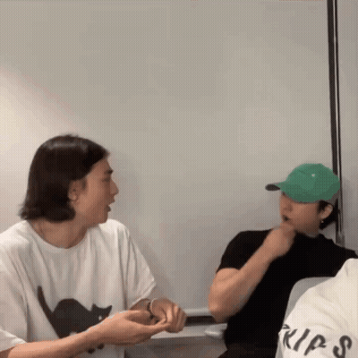 A Man Wearing A White Shirt With A Black Cat On It Is Talking To Another Man Wearing A Green Hat GIF