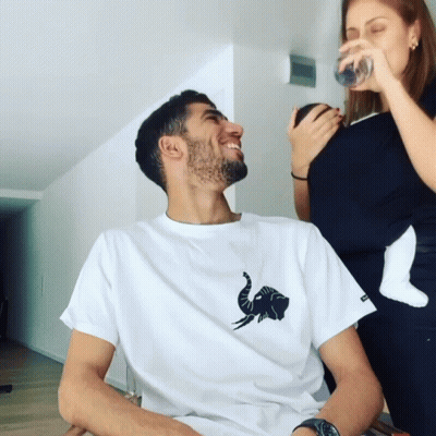 A Man Wearing A White Shirt With An Elephant On It Laughs While A Woman Drinks From A Glass GIF