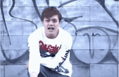 A Man Wearing A White Shirt With The Word Duck On It Stands In Front Of A Wall With Graffiti On It GIF