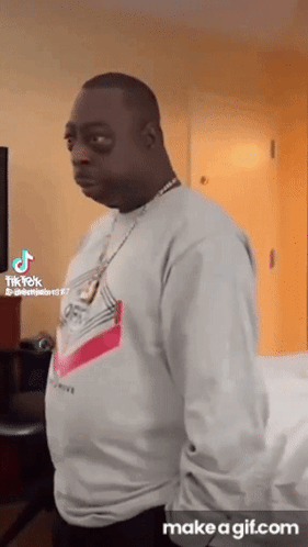 A Man Wearing A White Sweater And A Necklace Is Standing In A Room With His Hands In His Pockets . GIF