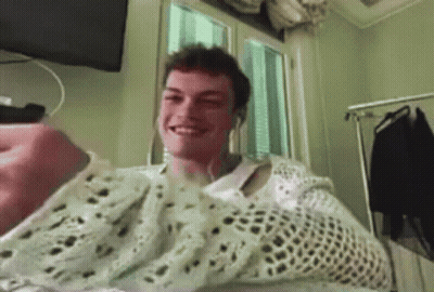 A Man Wearing A White Sweater And Headphones Is Smiling While Sitting In Front Of A Window . GIF