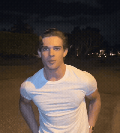 A Man Wearing A White T-Shirt And A Watch Stands In The Dark GIF