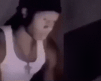 A Man Wearing A White Tank Top And A Black Hat Is Looking At A Laptop . GIF