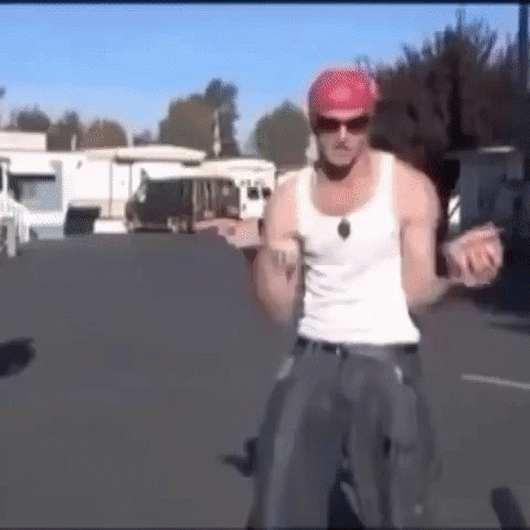 A Man Wearing A White Tank Top And A Red Hat Is Standing On A Bicycle . GIF