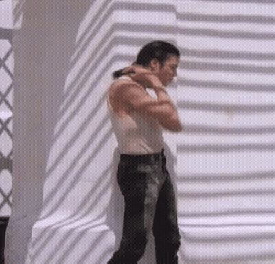 A Man Wearing A White Tank Top And Black Jeans Is Standing In Front Of A White Wall . GIF