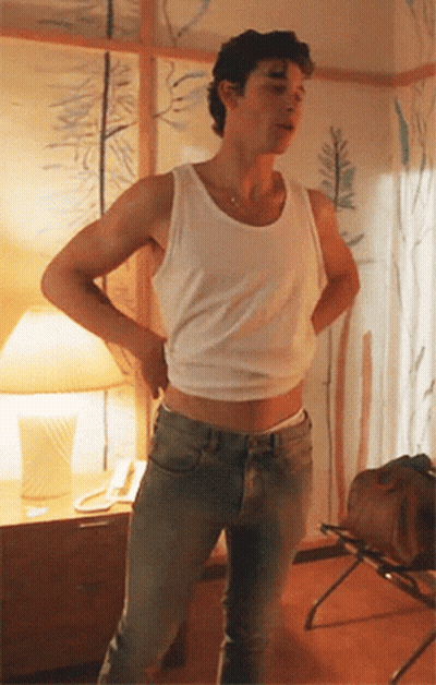 A Man Wearing A White Tank Top And Jeans Is Standing In A Room With His Hands On His Hips . GIF