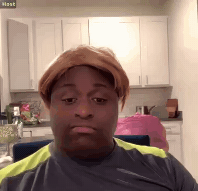 A Man Wearing A Wig Is Sitting In A Kitchen With A Sign That Says Host Above Him GIF