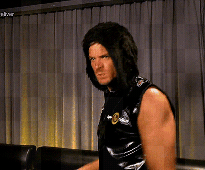 A Man Wearing A Wig Is Standing In Front Of A Curtain With The Word Eliver On The Bottom Right GIF