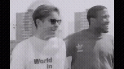 A Man Wearing A World In T-Shirt Is Standing Next To Another Man Wearing Sunglasses . GIF