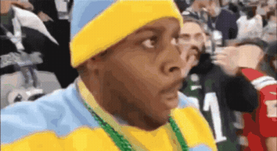 A Man Wearing A Yellow Hat And A Blue And Yellow Sweater With A Green Necklace Around His Neck GIF