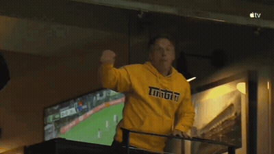 A Man Wearing A Yellow Hoodie With The Word Tigers On It Stands On A Balcony Watching A Soccer Game . GIF