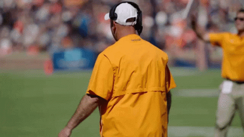 A Man Wearing A Yellow Jacket And A White Hat Stands On A Football Field GIF