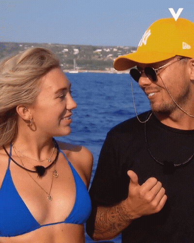A Man Wearing A Yellow Ny Hat Stands Next To A Woman In A Blue Bikini GIF