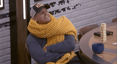 A Man Wearing A Yellow Scarf And A Hat Is Sitting At A Table GIF
