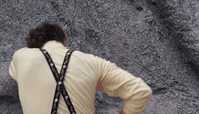 A Man Wearing A Yellow Shirt And Suspenders Is Standing In Front Of A Rock GIF