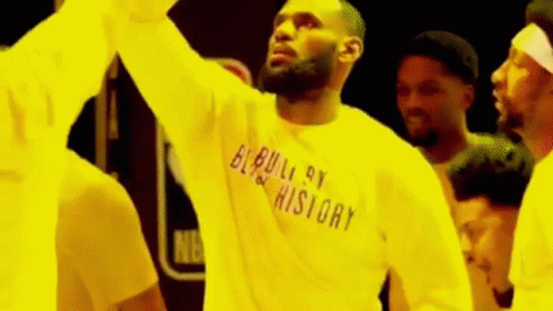 A Man Wearing A Yellow Shirt That Says `` Built By History '' Is Standing In Front Of A Crowd Of People . GIF