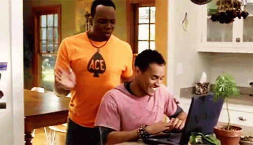 A Man Wearing An Ace Shirt Is Standing Next To Another Man Using A Laptop Computer GIF